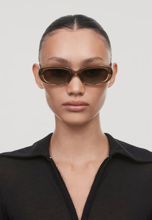 Lunettes de soleil rectangulaires marron avec une monture épaisse, dotées d'une finition brillante. Le modèle porte un haut noir avec des détails subtils autour du col.