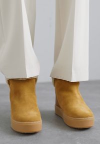 Yellow suede ankle boots with a textured finish, raised platform soles, and soft inner lining, paired with cream pants.
