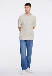 Camiseta de manga corta color beige claro con sutiles rayas horizontales, combinada con jeans de mezclilla azul y zapatillas blancas. Diseño simple y casual.