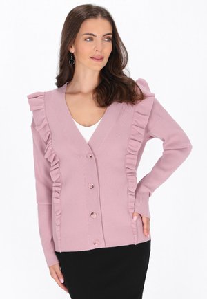 Frau trägt eine hellrosa, geknöpfte Strickjacke mit Rüschen-Details über einem weißen Top und einem schwarzen Rock und schaut zur Seite.