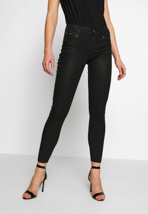 Frau trägt schwarze Skinny-Hosen und schwarze Sandalen mit hohen Absätzen und steht vor einem weißen Hintergrund.