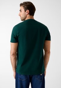 Zielona bawełniana koszulka z krótkimi rękawami, okrągłym dekoltem i prostym krojem, widziana od tyłu. Posiada subtelną metkę marki przy dolnym brzegu.