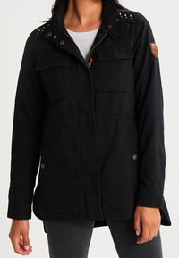 Černá pracovní bunda s hřebíkovaným límcem, dvěma předními kapsami a koženou nášivkou na rukávu; vyrobena z odolné látky s hladkým povrchem.