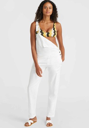 Witte katoenen overalls met verstelbare banden, een voorkant zak en accenten van zijknopen. Gekoppeld met een bikini-top met geometrisch patroon.