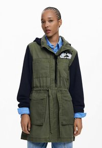 Zelena jakna od mješavine pamuka s crnom kapuljačom, elastičnim strukom, velikim prednjim džepovima i grafičkim printom na prsima. Stilirana s plavom košuljom ispod.