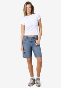 Denim šorti vidēji zili ar izplūdušu malu, pārī ar pieguļošu baltu t-kreklu un pelēkiem sporta apaviem. Modelis stāv neitrālā fona priekšā.