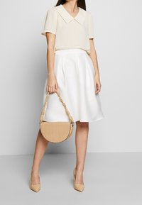 Blusa beige con un ampio colletto, gonna bianca e una borsa a mezzaluna in stampa coccodrillo color cammello con una tracolla intrecciata. Tacchi a punta color nude.