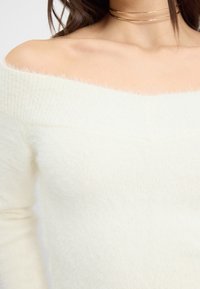 Maglione con scollatura a bardot, di colore crema, realizzato in un materiale morbido e soffice con texture a costine. Le caratteristiche principali includono un'ampia scollatura e maniche aderenti.