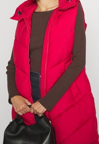 Rode puffer vest met hoge kraag en ritssluiting, voorzien van doorgestikte panelen. Draagt over een bruine geribde top met een zwarte handtas.
