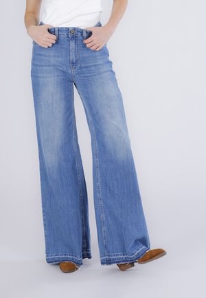 Persona che indossa jeans larghi a vita alta di colore azzurro chiaro e scarpe marroni, con le mani infilate nelle tasche anteriori, in piedi davanti a uno sfondo semplice.