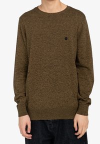 Brązowy sweter o fakturze z okrągłym dekoltem, długimi rękawami i małym czarnym logo na piersi. Nosi się go z ciemnymi dżinsami.