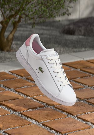 White leather sneaker with pink suede heel accent, lace-up front, green logo, flat sole, and minimalistic design, set on wooden tiles.
