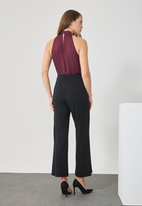 Haut halter bordeaux avec plis et dos en trou de serrure, associé à un pantalon large noir et des escarpins noirs, le tout sur un fond neutre.