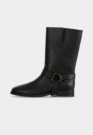 Botte noire en cuir mi-mollet avec bout pointu, talon bas et sangle décorative avec un détail d’anneau doré sur fond uni.