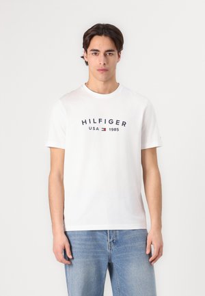 Bijela pamučna majica kratkih rukava s plavim natpisom "HILFIGER USA 1985". Glatka tekstura, okrugli izrez, opušteni kroj.