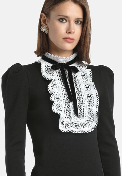 Schwarzes Kleid mit langen Ärmeln, versehen mit einem weißen Spitzenbesatz am Kragen und einem schwarzen Samtband. Texturiertes Gewebe mit gepufften Schultern.