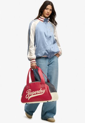 Mujer que lleva una chaqueta varsity azul y blanca y vaqueros, sosteniendo una bolsa de deporte Superdry roja y blanca por las asas.