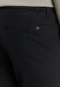 Gros plan d'un pantalon noir avec une poche passepoilée à l'arrière et un petit logo hexagonal au-dessus de la poche du côté droit.