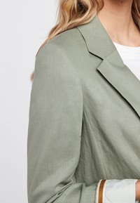 Blazer de lino verde con solapas de pico, costuras visibles y mangas con puños que presentan tela a rayas y un acento marrón contrastante.