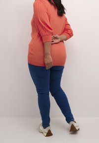 Femme en pull orange et jean bleu, les bras croisés dans le dos, debout contre un mur clair uni.