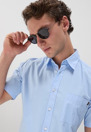 Giovane uomo con una camicia azzurro chiaro a maniche corte con bottoni che si aggiusta degli occhiali da sole neri rotondi.