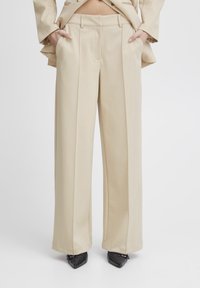 Beige wide-leg trousers with front pleats, smooth texture, and side pockets. Worn with pointed black shoes; no other details visible.