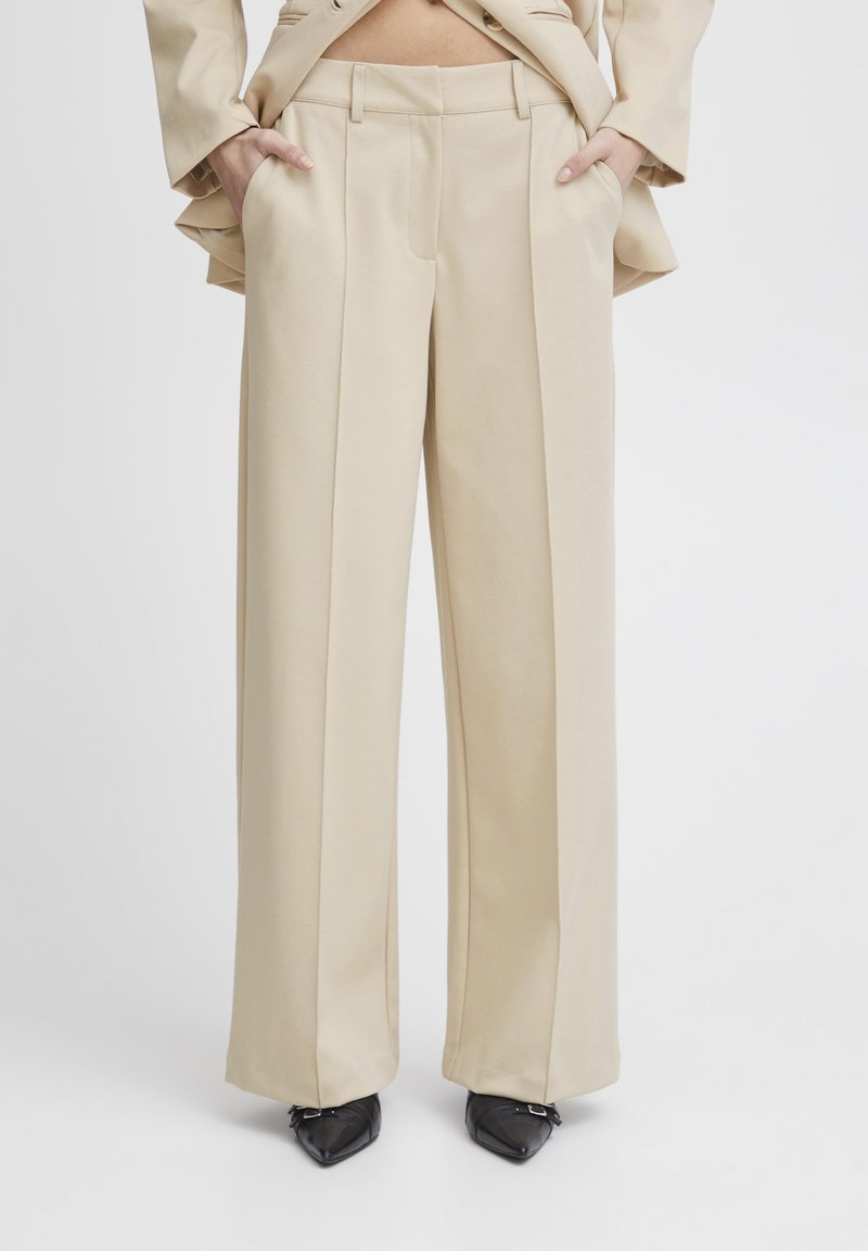 Beige wide-leg trousers with front pleats, smooth texture, and side pockets. Worn with pointed black shoes; no other details visible.