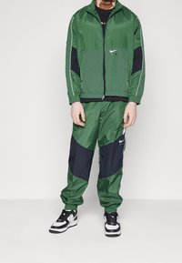 Completo sportivo in nylon verde con accenti neri, giacca con zip dotata di tasca frontale e pantaloni a sigaretta. Sneakers bianche e nere completano il look.