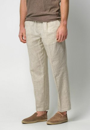 Lichtbeige linnenbroek met een getailleerde pasvorm, voorzien van een platte voorkant, plooien en een iets cropped lengte. Draagt erbij bruine instapschoenen.
