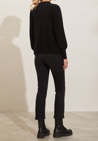 Pull en maille noir avec des manches bouffantes, ourlet en forme de scallop, associé à un jean noir et des bottes noires chunky. La tenue met en avant un design monochromatique.