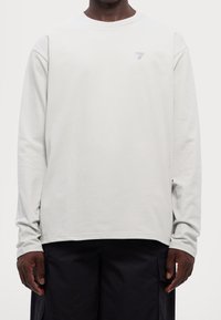 Hellgraues Langarmshirt mit einem strukturierten Muster und einem kleinen lila Logo mit der Nummer "7" auf der Brust. Der Stoff scheint weich und atmungsaktiv zu sein.