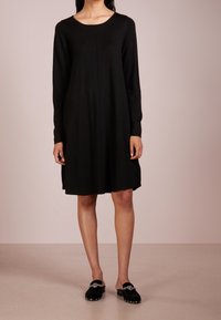 Schwarzes Langarmkleid mit rundem Ausschnitt, lockerer Passform und knielangem Saum. Kombiniert mit schwarzen Slippern mit dekorativen Akzenten.