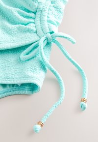 Gros plan d’un tissu texturé turquoise avec un cordon de serrage froncé noué en nœud, avec des perles métalliques dorées à chaque extrémité.