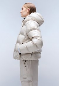 Giovane donna in una giacca puffer lucida di un leggero beige con cappuccio e pantaloni cargo coordinati, in piedi di profilo su uno sfondo semplice.