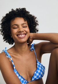Haut de bikini en vichy bleu avec des bords volantés, des liens ajustables et des accents en métal doré. Tissu doux, design léger, détail des épaules visible.