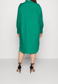 Grön linneskjortklänning med krage, lång ärm och avslappnad passform, kombinerad med svarta högklackade sandaler.