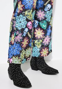 Stivaletti neri in suede con borchie argento, punta affusolata e tacco basso, abbinati a pantaloni colorati a fantasia floreale con disegni di teschi.