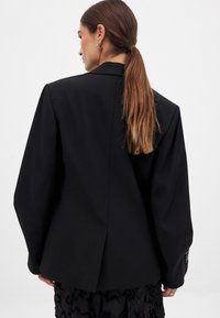 Sort blazer med strukturerede skuldre og en enkelt split bagpå. Skræddersyet design med tekstureret stof og knapdetaljer på manchetterne.