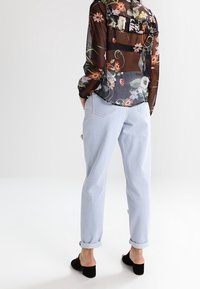 Blouse noire transparente à fleurs avec volants, associée à un pantalon bleu clair à revers. Des chaussures noires à enfiler complètent la tenue.