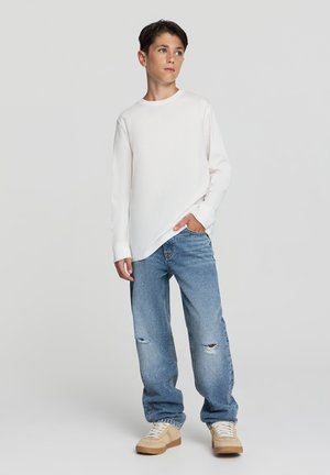 Camicia bianca a maniche lunghe, jeans strappati blu e sneakers beige; vestibilità comoda con un aspetto casual. Design semplice, texture morbida.