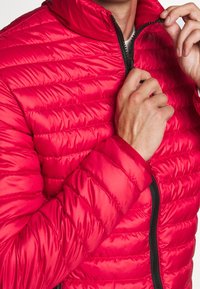 Červená péřová bunda s horizontálně prošívanými sekcemi, vysokým límcem a černými zipovými detaily. Materiál vypadá leskle a má hladkou texturu.