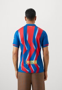 Mėlyna ir raudona polo marškinėliai su zigzaguota raštais, tekstūruotas audinys, trumpomis rankovėmis ir siuvinėtu logotipu apatinėje priekyje.