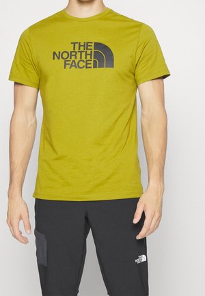 Olivengrünes Baumwoll-T-Shirt mit kurzen Ärmeln, rundem Halsausschnitt und einem großen schwarzen Logo auf der Brust. Trägt eine dunkle Hose.