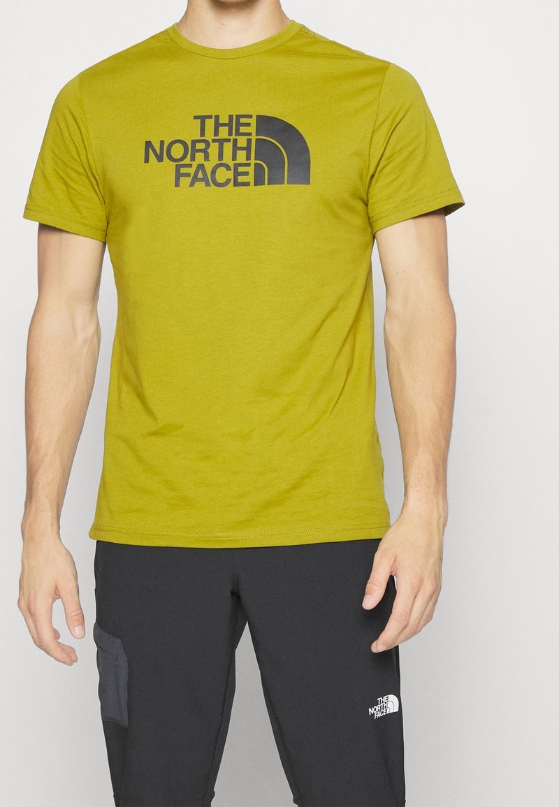 Olijfgroene katoenen T-shirt met korte mouwen, ronde halslijn en een groot zwart logo over de borst. Draagt met donkere broek.