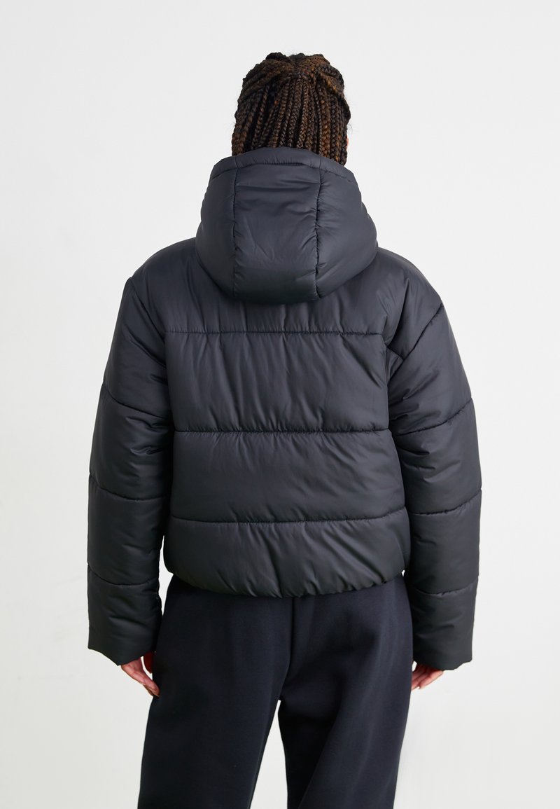 Giacca nera e gonfiabile con cappuccio, caratterizzata da un design trapuntato e polsini elasticizzati, abbinata a pantaloni scuri. Texture liscia, stile minimalista.