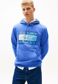 Junger Mann trägt einen blauen Tommy Jeans Hoodie mit einer Fronttasche, kombiniert mit blauen Jeans und einem weißen Shirt darunter.