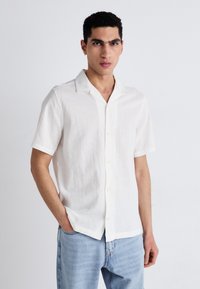 Chemise en lin blanche à manches courtes avec une texture douce, col classique et boutons jaunes, associée à un jean en denim bleu clair.