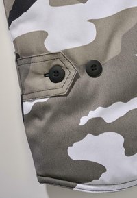 Tela de camuflaje en tonos de gris y blanco, con una solapa de bolsillo hexagonal y dos botones negros. Textura suave.