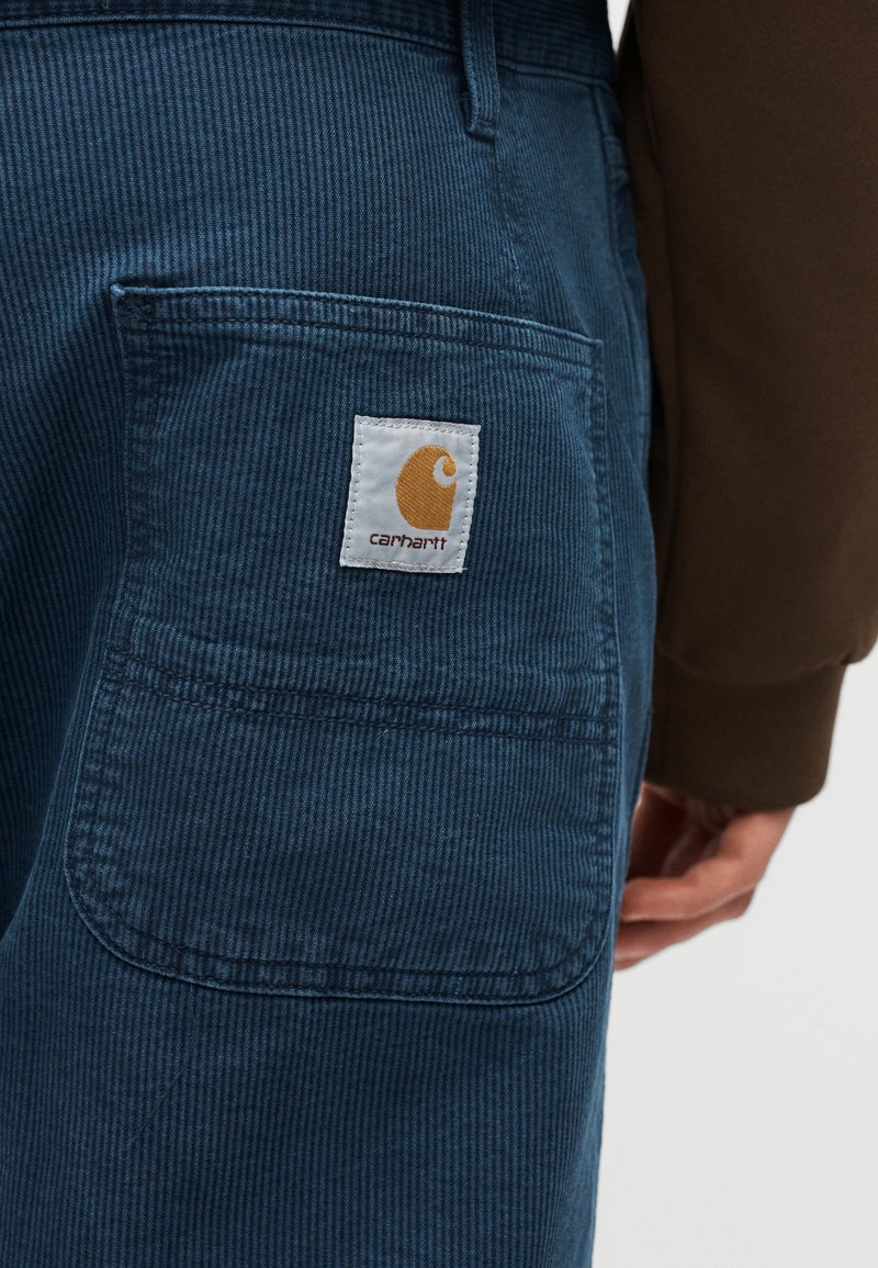 Pantalon en velours côtelé bleu avec une poche arrière visible ornée d'un patch logo Carhartt, porté par une personne portant une chemise marron à manches longues.