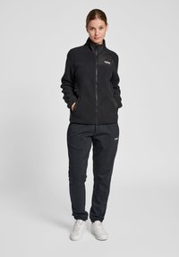 Svart fleecejacka med dragkedja och hög krage, tillsammans med svarta sweatpants. Båda har fickor, och jackan har en logga på bröstet.